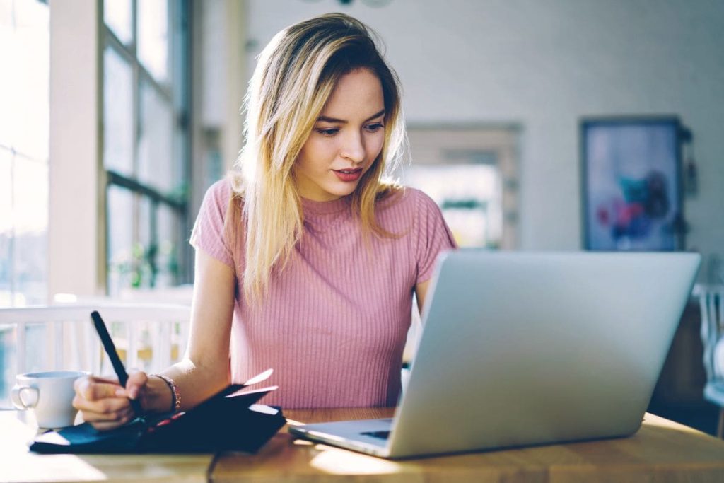 A Content Moderator working remotely from home, using a laptop and writing notes in a notebook.