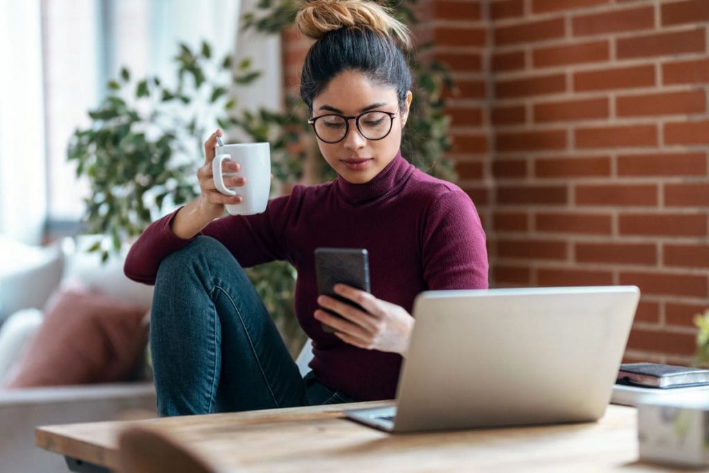 17 Easy Passive Income Ideas for Beginners Woman sitting at her home office desk, using her phone while drinking coffee.