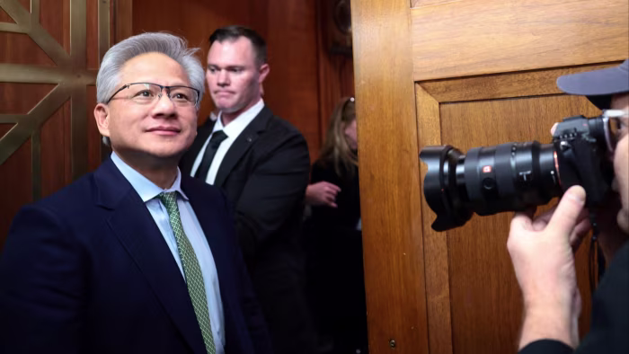 Nvidia can sell H200 AI chips to China, Donald Trump says Jensen Huang stands near a wooden door, looking ahead as a photographer takes his picture. A security guard is visible behind him.