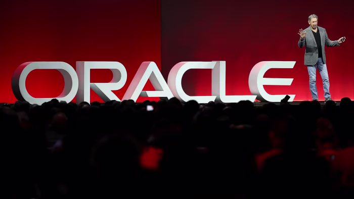 Oracle shares slide as it adds $15bn to data centre spending Larry Ellison stands on stage in front of a large ‘ORACLE’ sign, delivering a keynote to an audience.