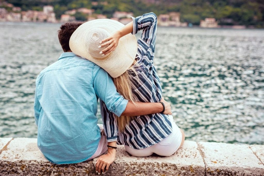 24 Romantic Business Ideas That Let You Profit From Love A couple sitting together on a rock wall, overlooking a lake with hills and buildings in the distance.
