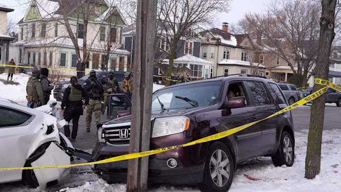 ICE officer fatally shoots Minnesota woman in her car Federal agents in tactical gear stand near a maroon SUV with a bullet hole in the windshield and a damaged white car, behind yellow police tape.