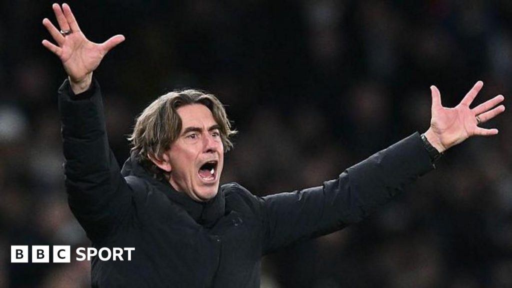 Tottenham head coach Thomas Frank, in a black coat, has his arms and hands outstretched in an animated gesture