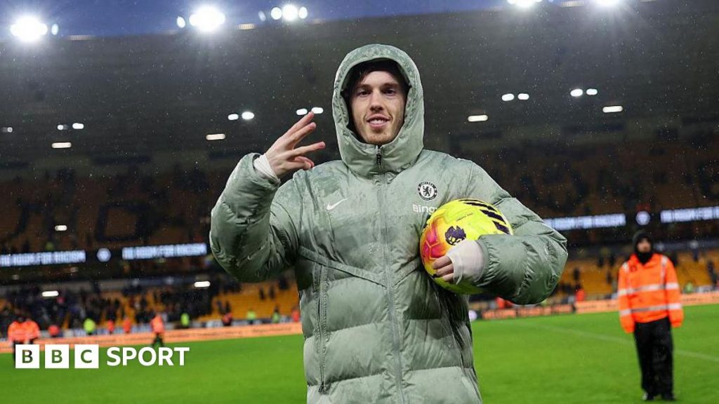 Cole Palmer celebrates scoring a hat-trick for Chelsea with a three-finger salute with his right hand, while holding the yellow match ball under his left arm and wearing a large Chelsea puffer jacket with the hood up.