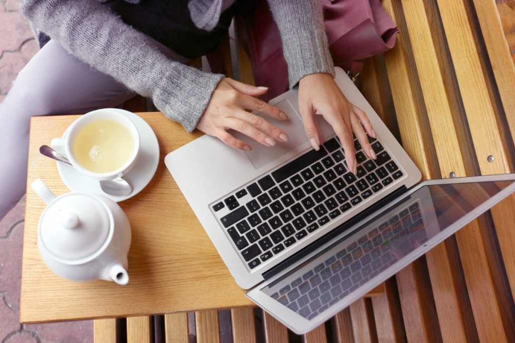 12 Remote Jobs With Little Human Interaction A closeup image of a woman's hands typing on a laptop while working remotely and drinking tea.