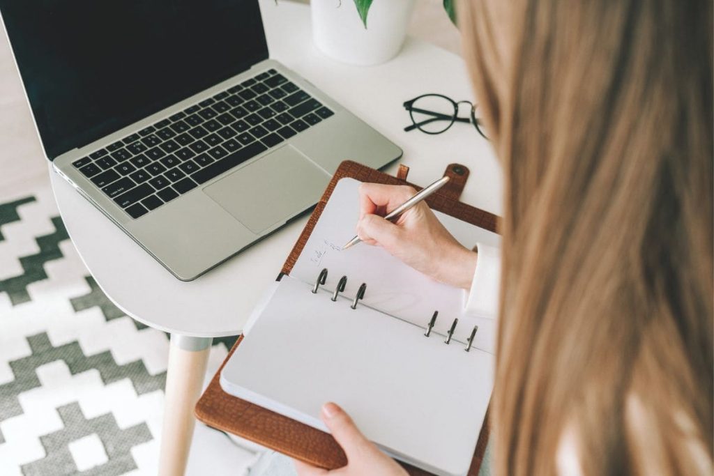 A woman writes in a notebook while using a laptop to job hunt and create a resume.