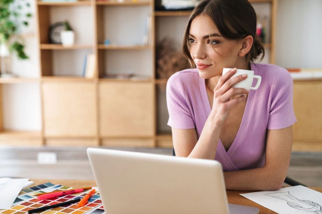 A woman working from home, sitting at a desk, using a laptop, and drinking coffee.