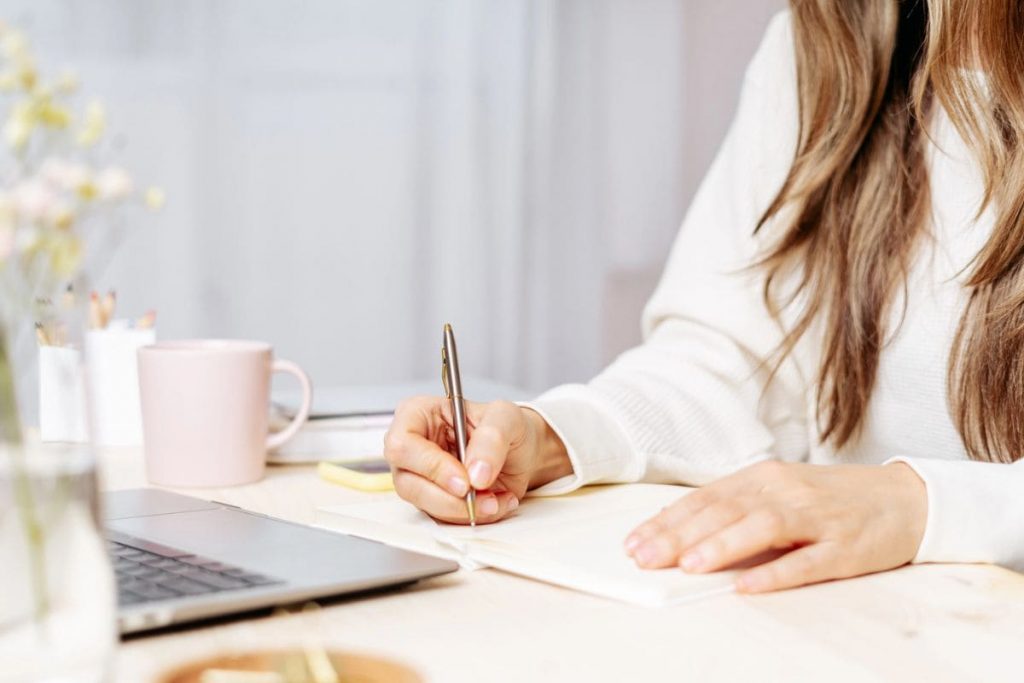 A woman working from her home office desk, using a laptop and writing in a notebook.