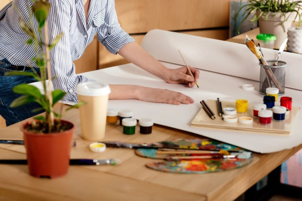A closeup view of an artist's desk: a woman is using paints to color a large canvas.