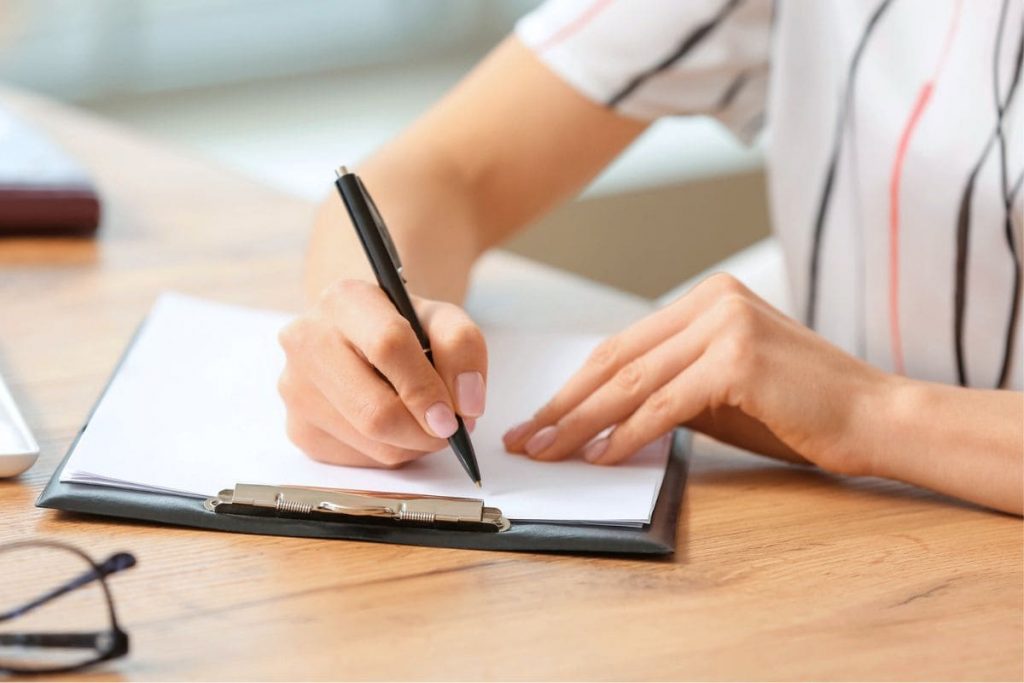 A woman sitting at a desk, writing notes on a clipboard.