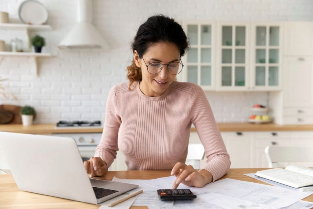 A woman working remotely from her kitchen table, using a laptop and a calculator.