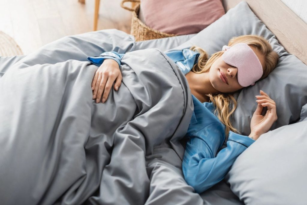 A woman sleeping in bed and wearing a sleep mask for blog post get paid to sleep.