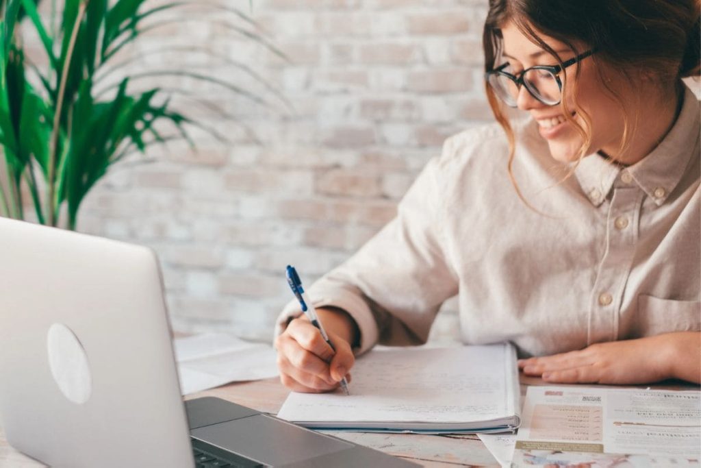 9 Top AI Training Sites to Earn Extra Cash From Home A woman working remotely from her home office, using a laptop and writing in a notebook.
