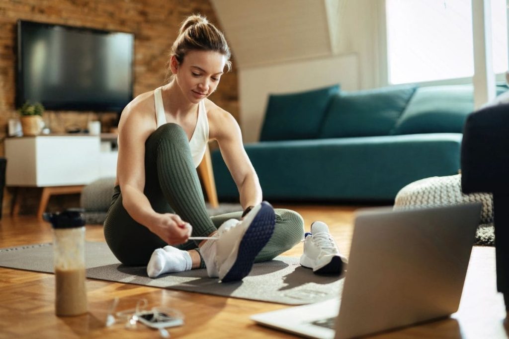 Make Money & Get Fit A woman getting ready to exercise at home, sitting in her living room on a yoga mat, putting on running shoes and using a laptop.