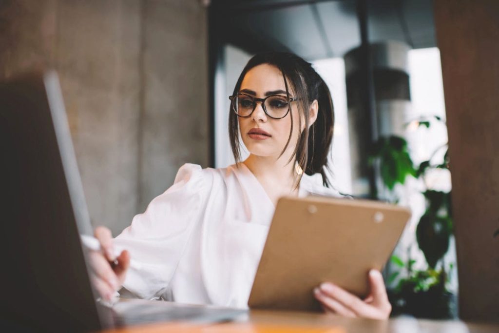 A recruiter sitting at a desk, holding a clipboard, looking at a resume, while using laptop.