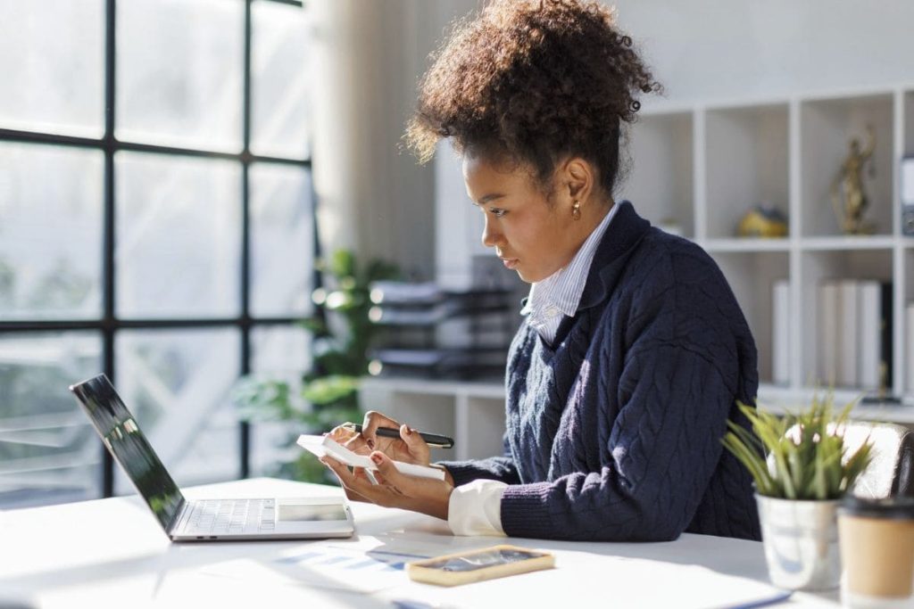A woman accountant, sitting in her home office, using calculator while working at her computer.