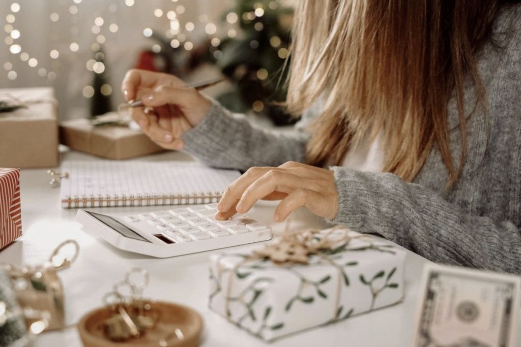 A woman creating a holiday budget, using a calculator and writing in a notebook while surrounded by holiday decor.